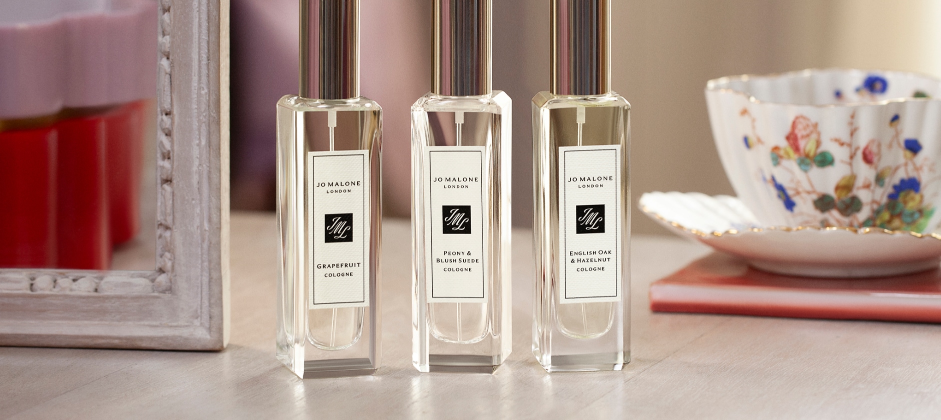 Three Jo Malone London perfume bottles with silver caps on a wooden table, set beside a decorative teacup and a framed mirror